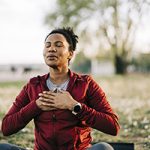 African American woman taking deep relaxing breaths while sitting cross-legged outdoors.