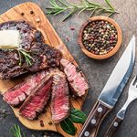 Medium rare Ribeye steak with herbs and a piece of butter on the wooden tray. A steak knife and fork rest nearby, along with a small wooden bowl full of peppercorns and sprigs or rosemary and thyme.