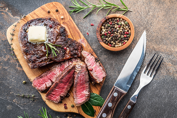 Medium rare Ribeye steak with herbs and a piece of butter on the wooden tray. A steak knife and fork rest nearby, along with a small wooden bowl full of peppercorns and sprigs or rosemary and thyme.