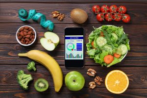 Smartphone with weight loss calculator application, measuring tape and different products on wooden table, flat lay