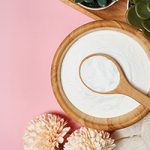 Collagen powder in a wooden bowl and spoon on pink table, surrounded by plants.