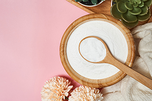 Collagen powder in a wooden bowl and spoon on pink table, surrounded by plants.