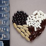 Supplements and tablets laid out in heart shapes next to the pillbox by day on a gray background