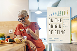 Background photo of mature woman with short grey hair and a red t-shirt, leaning against a wooden countertop as she eats a large salad from a clear bowl. On top, book cover of "The Origin of Being" by Luke Comer and Jenny Powers, PhD.