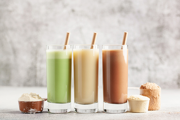 A trio of protein shakes with straws, along with measuring scoops of protein powders.