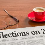 A newspaper on a wooden desk with he headline reading, "Reflections on 2024." A pair of glasses and cup of espresso sit nearby.