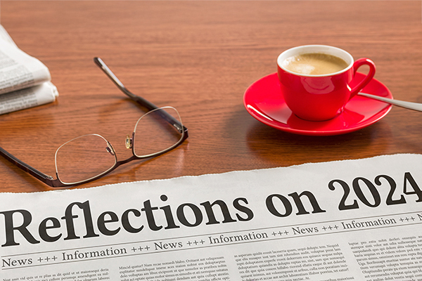 A newspaper on a wooden desk with he headline reading, "Reflections on 2024." A pair of glasses and cup of espresso sit nearby.