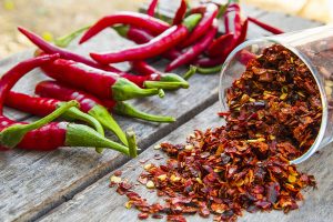 A wooden table background, with a bunch of red chili peppers and small bowl spilling red pepper flakes.