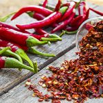 A wooden table background, with a bunch of red chili peppers and small bowl spilling red pepper flakes.