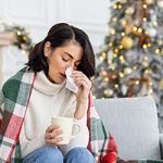 A woman draped in a blanket holding a cup of tea and wiping her nose. In the background, a Christmas tree is visible.