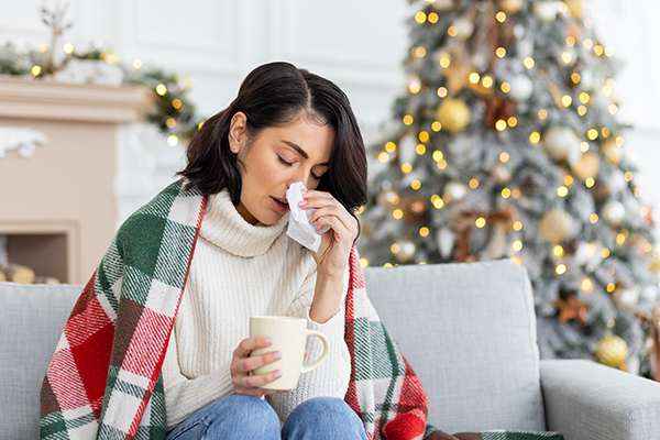 A woman draped in a blanket holding a cup of tea and wiping her nose. In the background, a Christmas tree is visible.