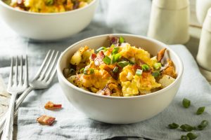 In the foreground, a white bowl filled with an egg, potato and bacon scramble. A fork rests nearby, and part of a second bowl can be seen in the top left corner of the image.