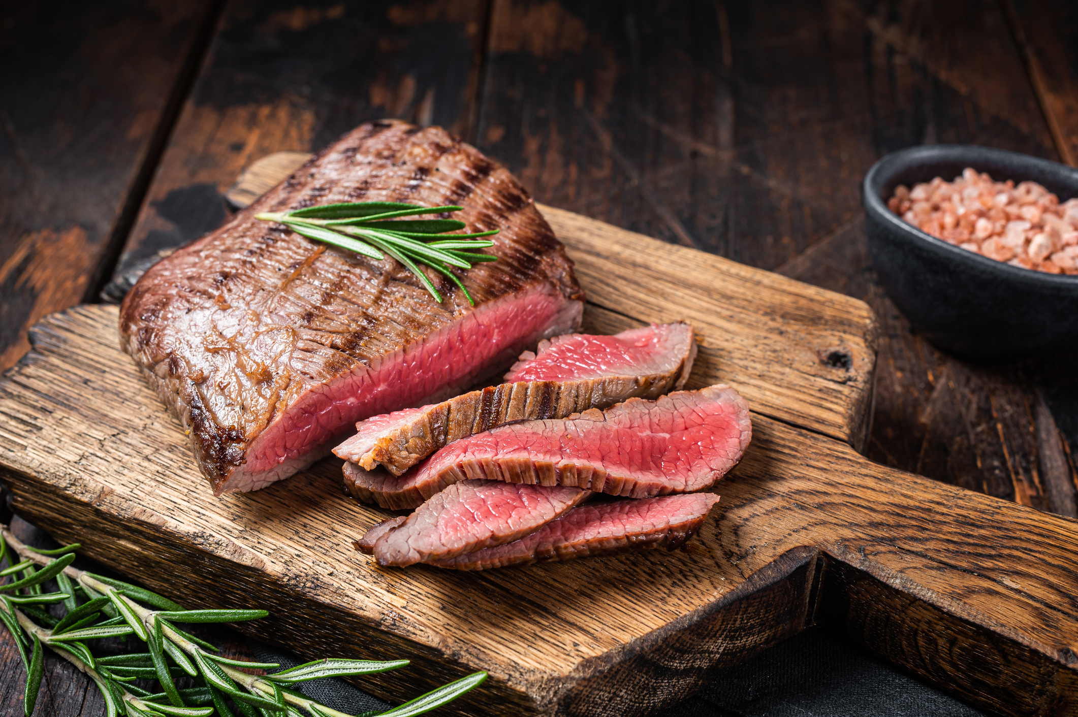 Barbecue dry aged wagyu Flank Steak on a cutting board, garnished with sprigs of rosemary. Wooden background.