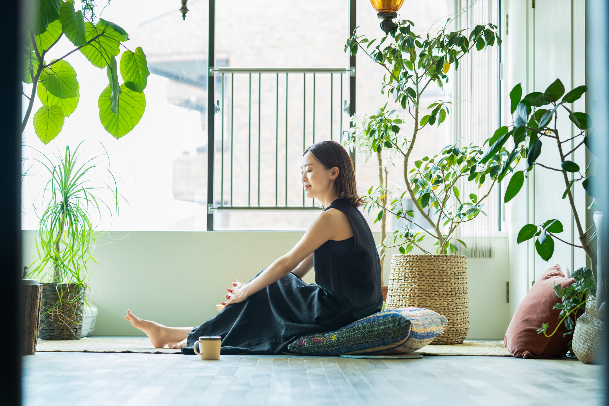 A woman sitting on a cushion in a sun-drenched room with house plants around her. There's a mug sitting on the floor near her, and she looks peaceful and relaxed.