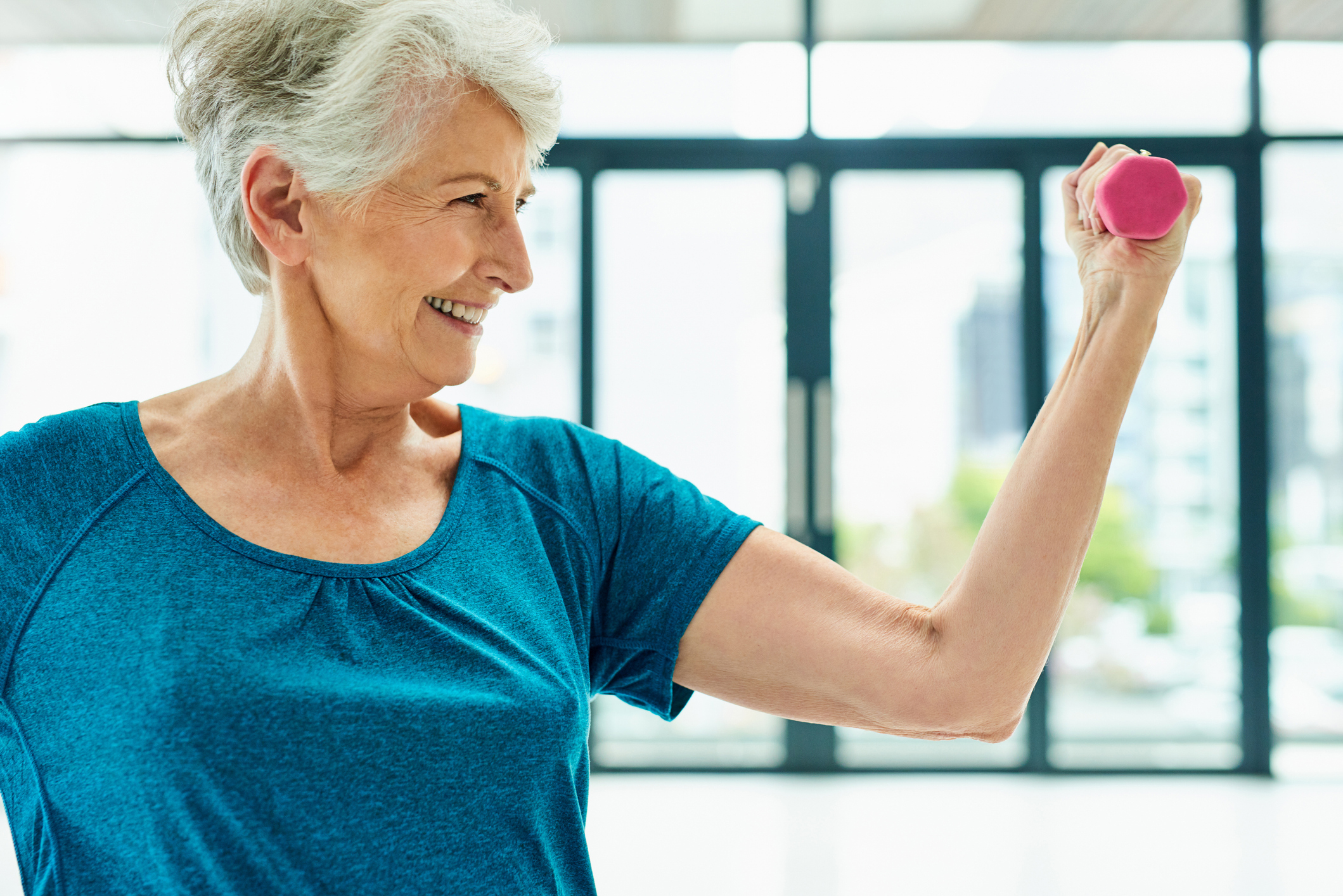 Senior woman with short hair dressed in a blue t-shirt, flexing her arm as she lifts a lightweight dumbbell