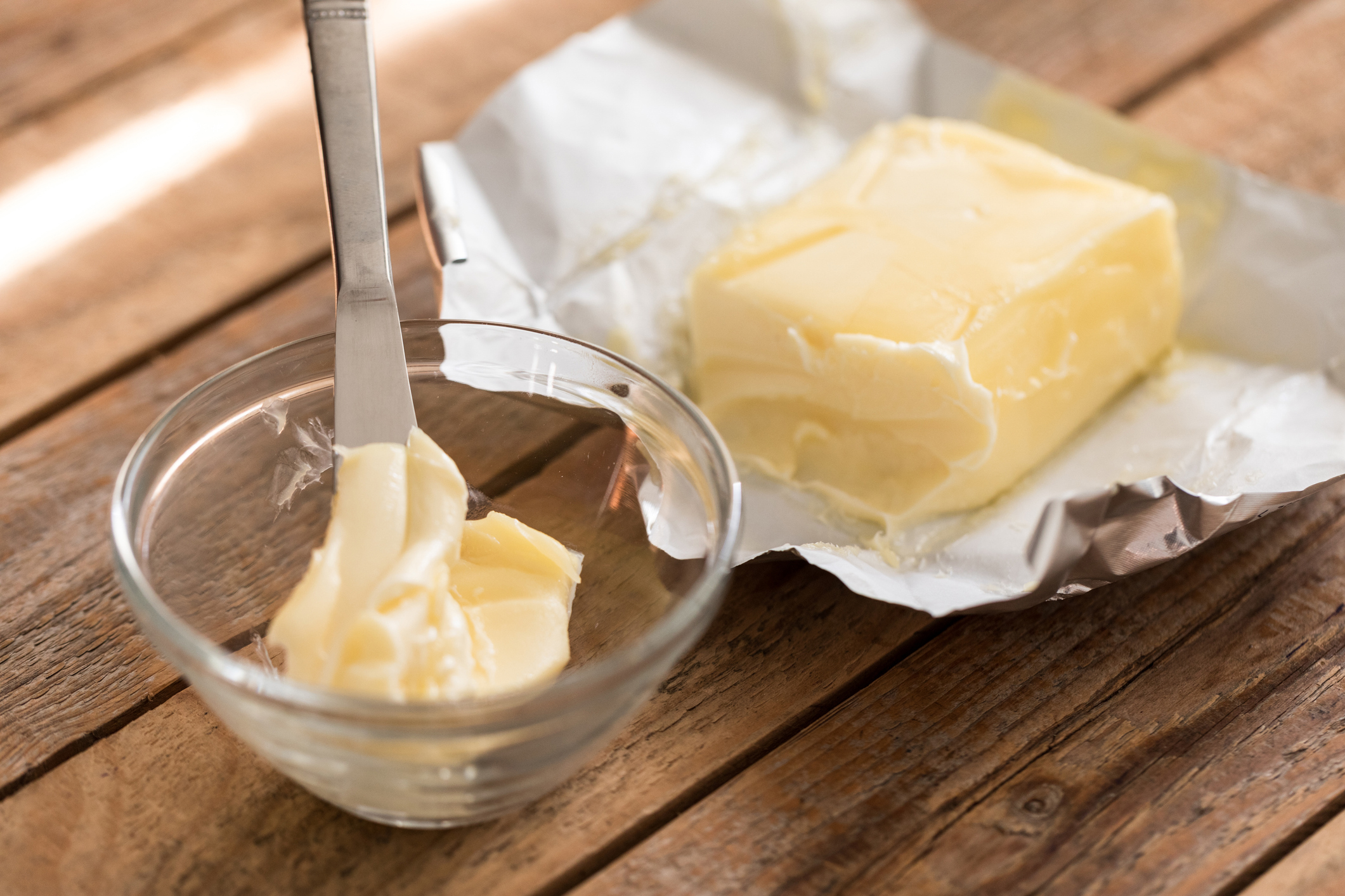 One pound brick of butter in its wrapping with some butter scooped off, resting on a wooden table. Next to it, a small glass bowl holds the butter that's been scraped from the brick and a butter knife.