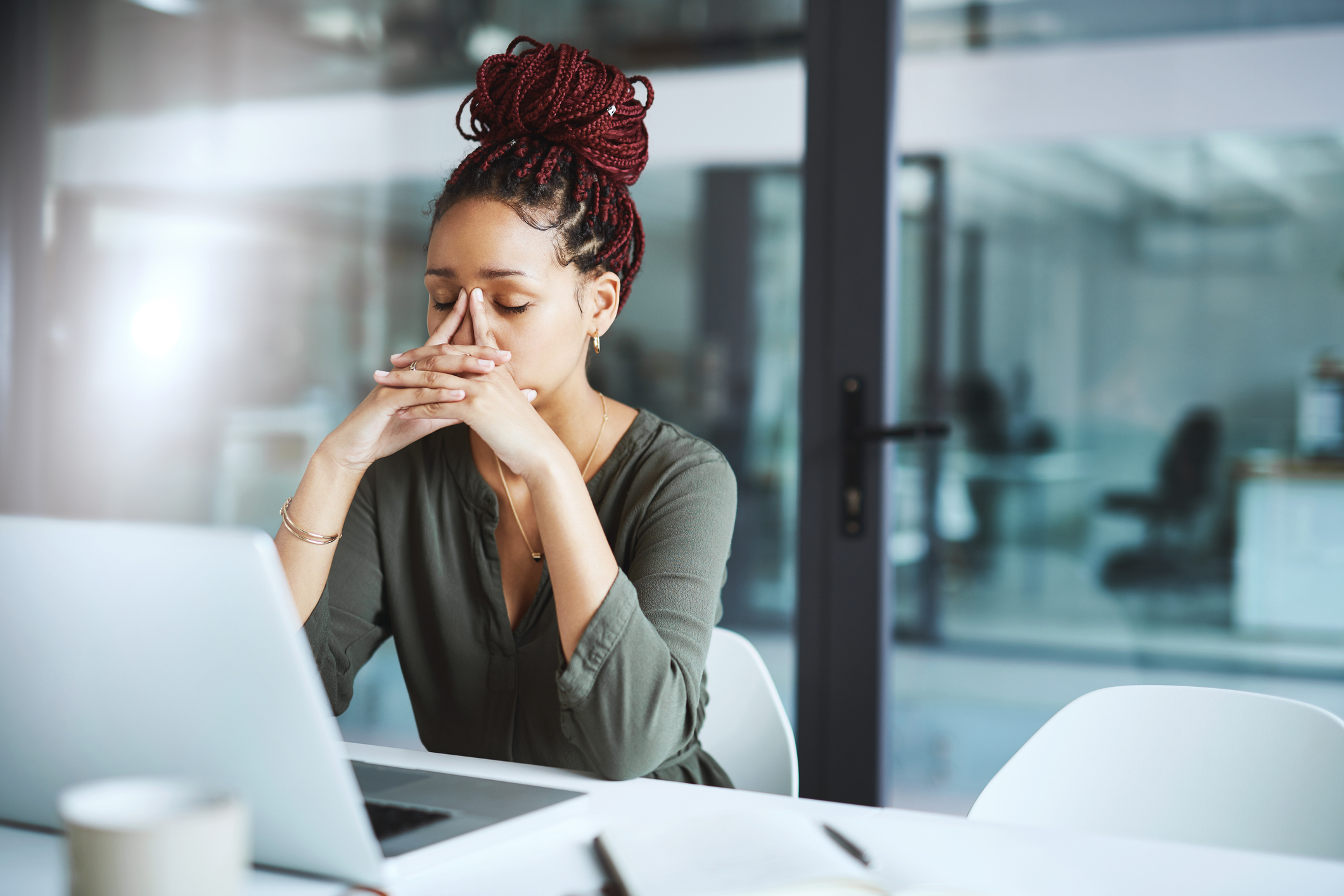 African American woman sitting at her desk in front of her open laptop. She has her elbows resting on the table and her fingers steepled against her face, eyes closed as she takes a moment in the stress of her workday.