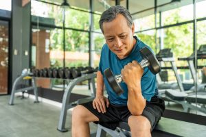 Mature Asian man sitting on a weight bench and doing bicep curls with a dumbbell.