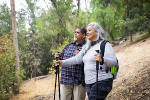 Mature Mexican couple enjoying the view during a hike through the woods. They both carry hiking poles and wear backpacks. He is in a plaid shirt and light colored pants, while she wears a gray long-sleeved top and patterned leggings.