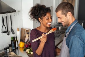 Multiethnic couple tasting food from wooden spoon while standing in their kitchen