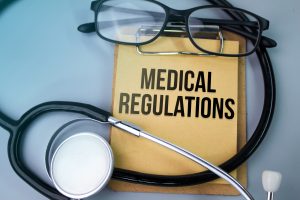 Stethoscope and glasses resting over s clipboard with yellow paper. On the paper is printed the words "medical regulations."