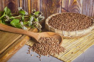 A woven basket filled with groats of buckwheat, alongside wooden cutting board on which rests a blooming branch buckwheat and wooden spoon with buckwheat groats spilling from it