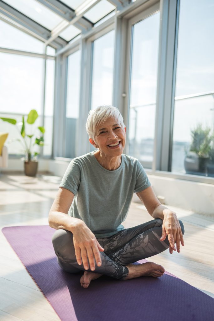 A cheerful older woman enjoys a moment of relaxation, practicing gentle exercises on a yoga mat within a bright space surrounded by large windows, promoting positivity and peace.