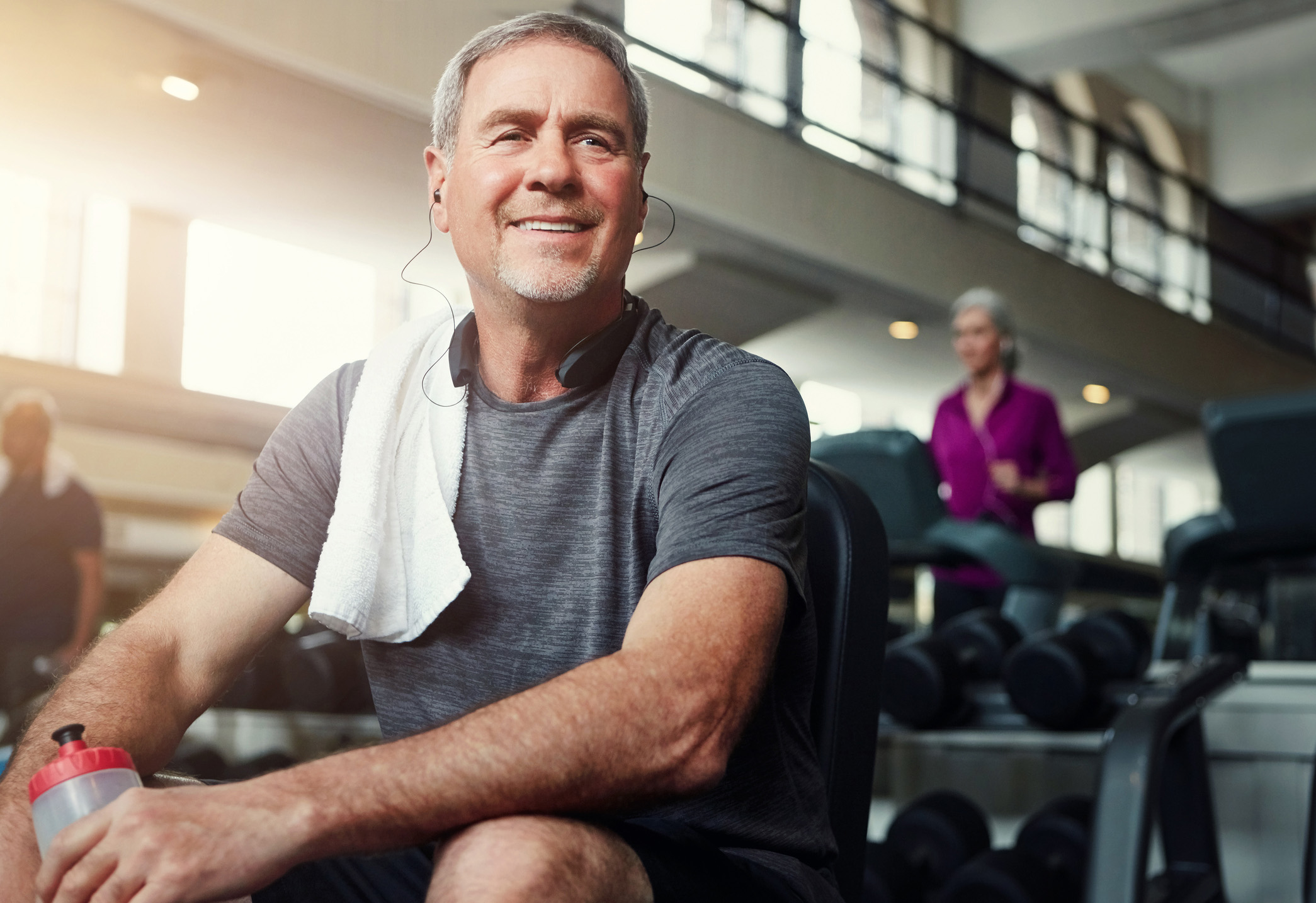 Middle aged man sitting on a weight bench with a towel over his shoulder and earbuds in his ears is taking a break from his workout