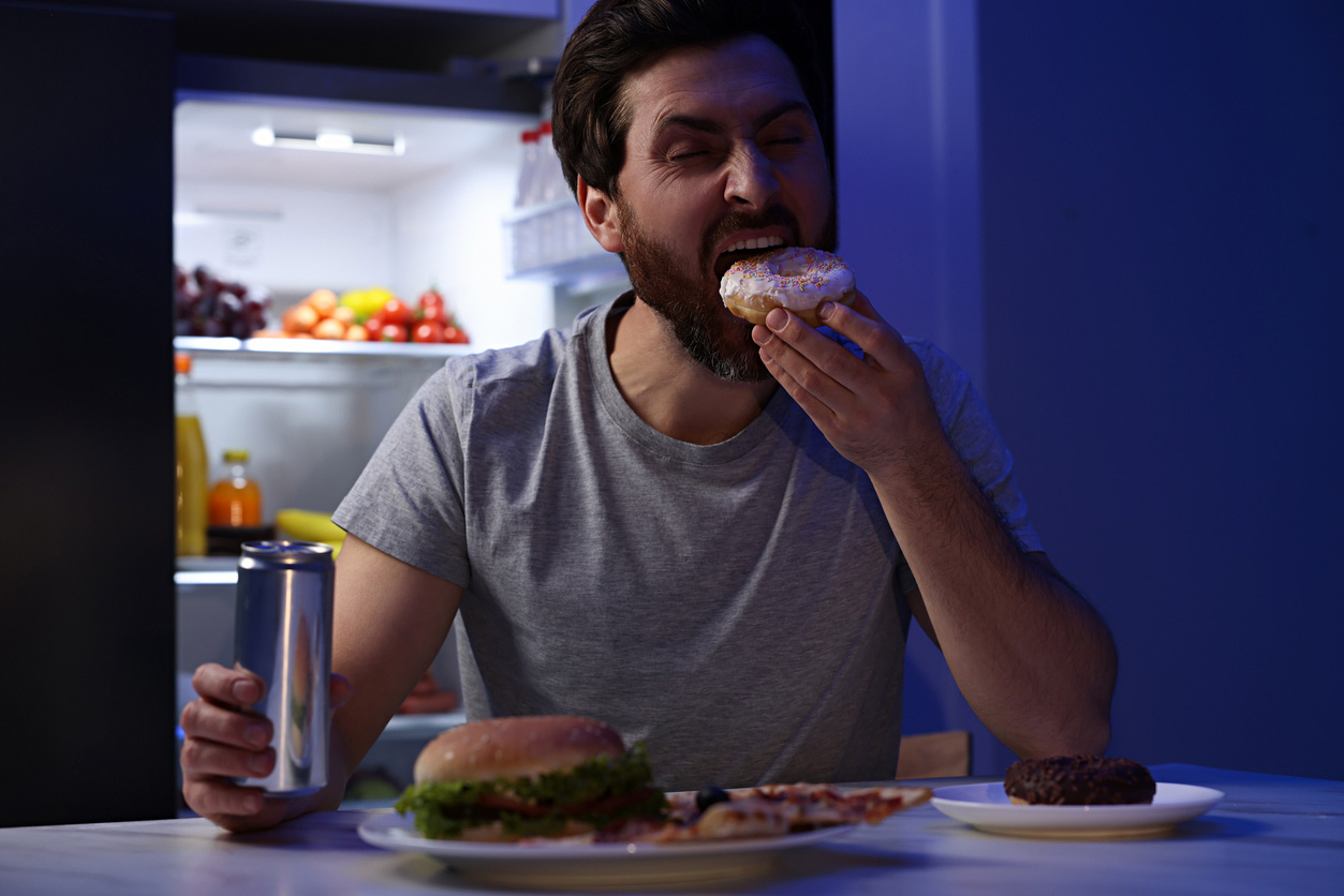 Man holding tin can and eating donut in kitchen at night, with an open fridge behind him and a burger on a plate in front of him.