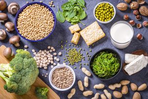 Top view of various kinds of vegan protein sources like tofu, tempeh, soy beans, soy milk, mushrooms, wakame, lentils, peanuts, spinach and chick peas. All the objects are on a gray bluish backdrop.