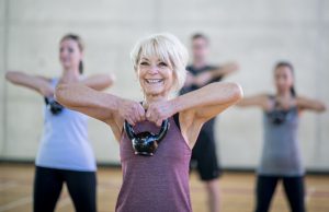 An older Caucasian woman is seen doing a squat with a kettlebell, while participating in a co-ed, multi-ethnic, fitness class.