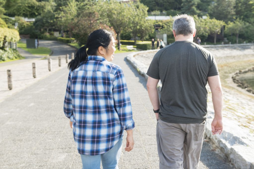 This is a horizontal, color photograph of a happily married mixed race Canadian couple spending the day together walking along False Creek Bay in downtown Vancouver, Canada. They walk side by side away from the camera.