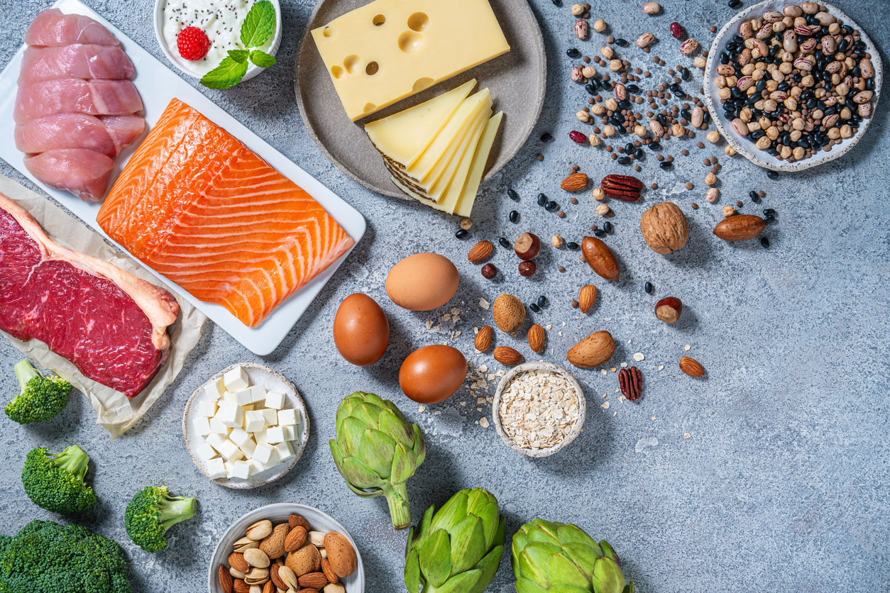 A variety of nutritious protein sources are displayed on a gray kitchen table. Ingredients include salmon, chicken, beef, eggs, cheese, and legumes, along with vegetables.