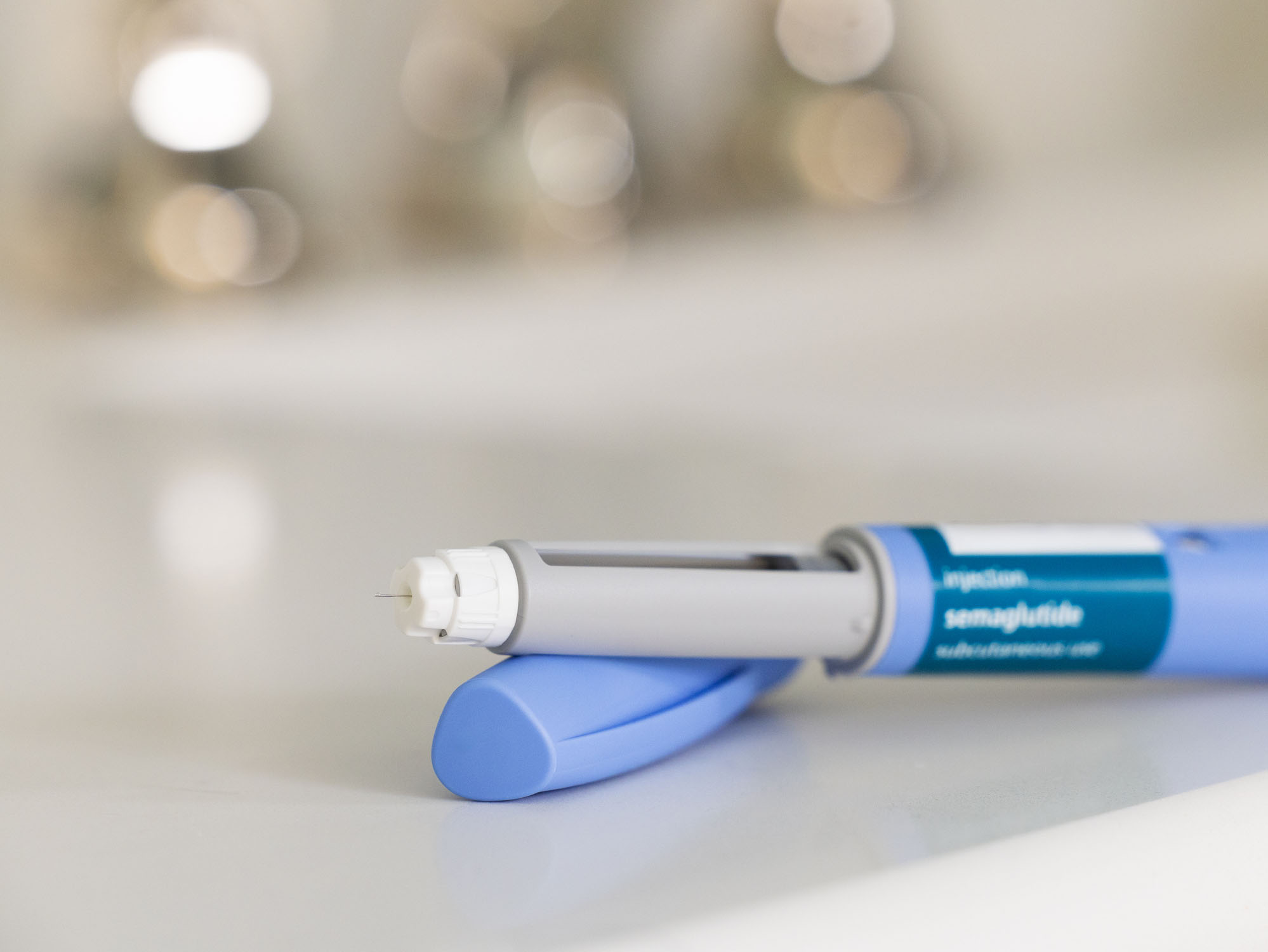 GLP-1 semaglutide injection pen, a medication used to treat Type 2 diabetes and aid in weight loss, on a white surface. The prescription medicine is used to treat Type 2 diabetes as It helps regulate blood sugar levels by stimulating insulin secretion and decreasing glucagon levels. In addition, it is also used off-label for weight loss in individuals with obesity, as it can help reduce appetite and increase feelings of fullness. The pen is positioned horizontally, with its cap removed, on a bathroom countertop with a white surface. The medical device is in the foreground and the background is out of focus because of a shallow depth of field.