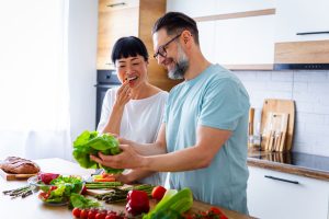 Cheerful couple preparing fresh vegetables in a modern kitchen. A variety of colorful vegetables, a bowl, and a loaf of bread are placed on the wooden countertop. They are smiling and enjoying the cooking process together.