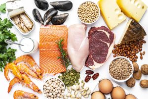 Overhead view of a large group of food with high content of healthy proteins. The composition includes salmon beef fillet, chicken breast, eggs, yogurt, mussels, chick peas, pistachios, cheese, brown lentild, beans, shrimps, canned tuna, pumpkin seeds, soybeans among others.