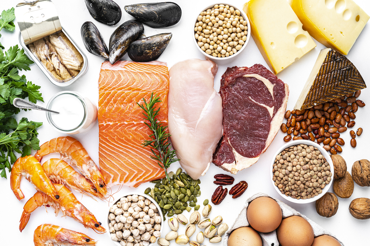 Overhead view of a large group of food with high content of healthy proteins. The composition includes salmon beef fillet, chicken breast, eggs, yogurt, mussels, chick peas, pistachios, cheese, brown lentild, beans, shrimps, canned tuna, pumpkin seeds, soybeans among others.
